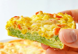 Fatia de torta de brócolis saudável sendo segurada pela mão, mostrando o recheio suculento com milho, pimentão e queijo derretido