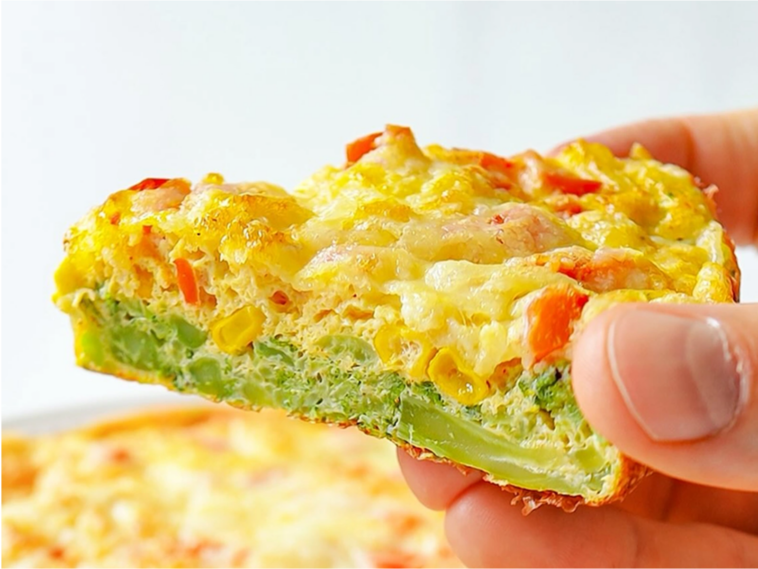 Fatia de torta de brócolis saudável sendo segurada pela mão, mostrando o recheio suculento com milho, pimentão e queijo derretido